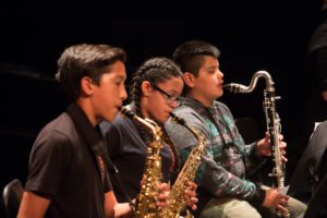 three students play wood wind instruments. two students play saxophone and one plays bass clarinet