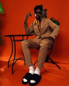 Henry posing sitting in a chair in a tan suit, orange background